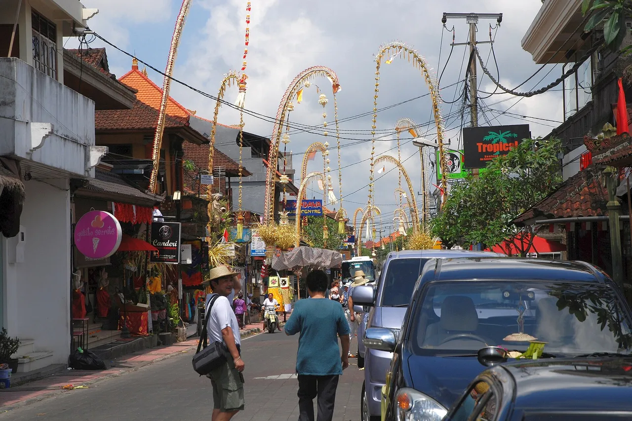 A shopping street in Ubud decorated with penjor poles, cars at a standstill and pedestrians walking on the roadway, Bali