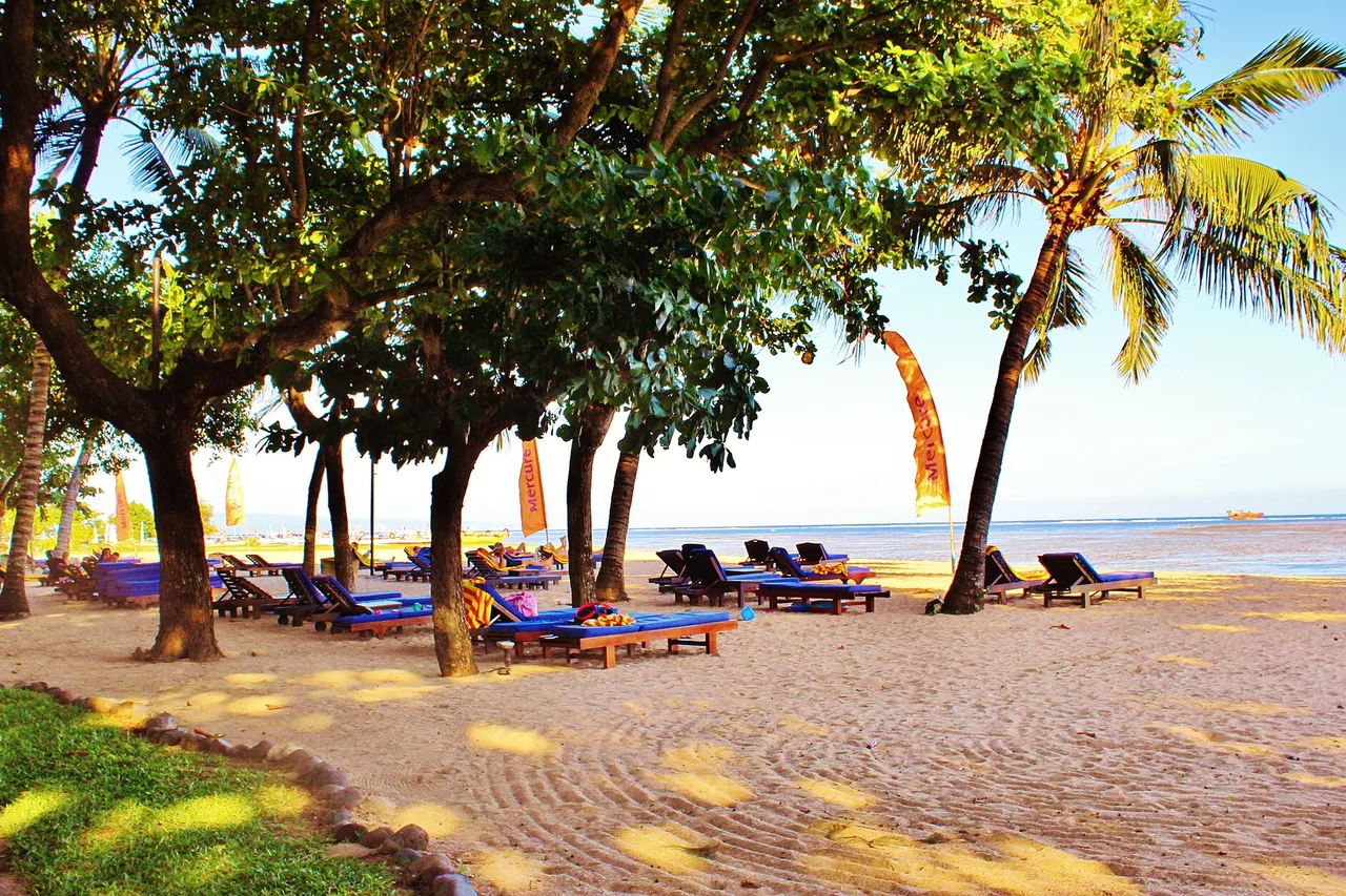 Loungers under the trees and penjor poles planted in the sand, the calm lagoon at Sanur, Bali