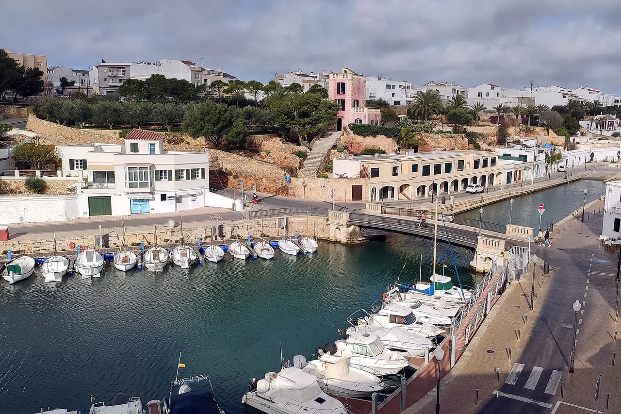 Der Hafen von Ciutadella auf Menorca von oben: ein schmales Becken mit vertäuten Booten, die niedrigen Häuser am Kai und die Straße am Wasser entlang.