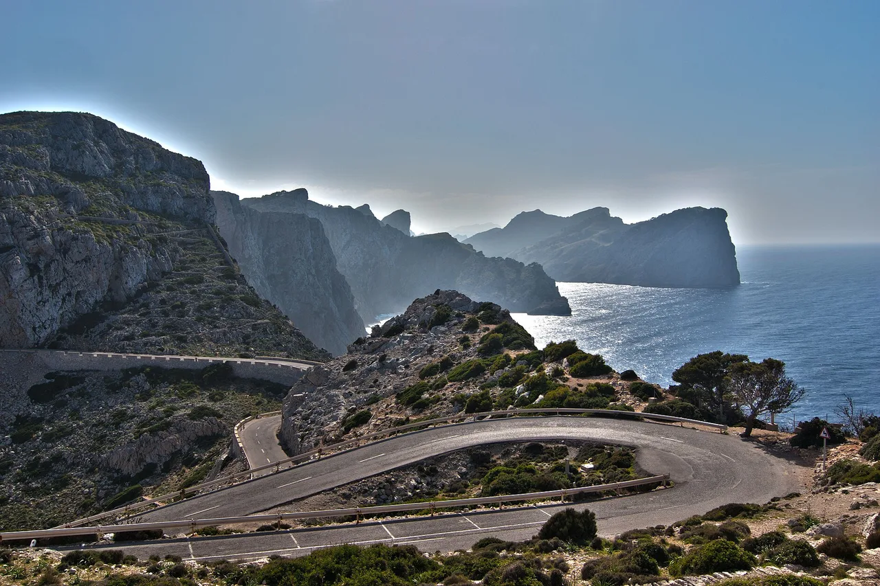 Die Serpentinenstraße zum Cap de Formentor auf Mallorca: die Asphaltschleife im Vordergrund, dahinter Klippen und Meer, und das Kap, das im Dunst verschwindet.