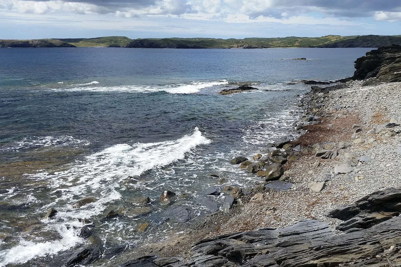 La côte d’ardoise noire près du phare de Favàritx : des dalles sombres et feuilletées plongeant dans une mer agitée qui déferle en écume, une bande de galets clairs, et à l’horizon les collines vertes de la côte nord.