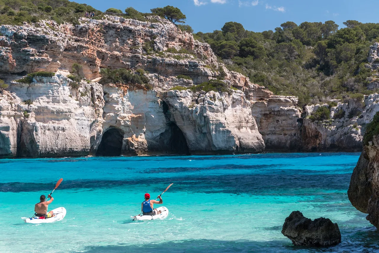 Deux kayakistes pagaient sur l’eau turquoise très claire d’une crique de Minorque, devant une falaise calcaire blanche creusée de grottes ; en haut de la falaise, quelques promeneurs minuscules sur le belvédère, et une pinède en bordure.