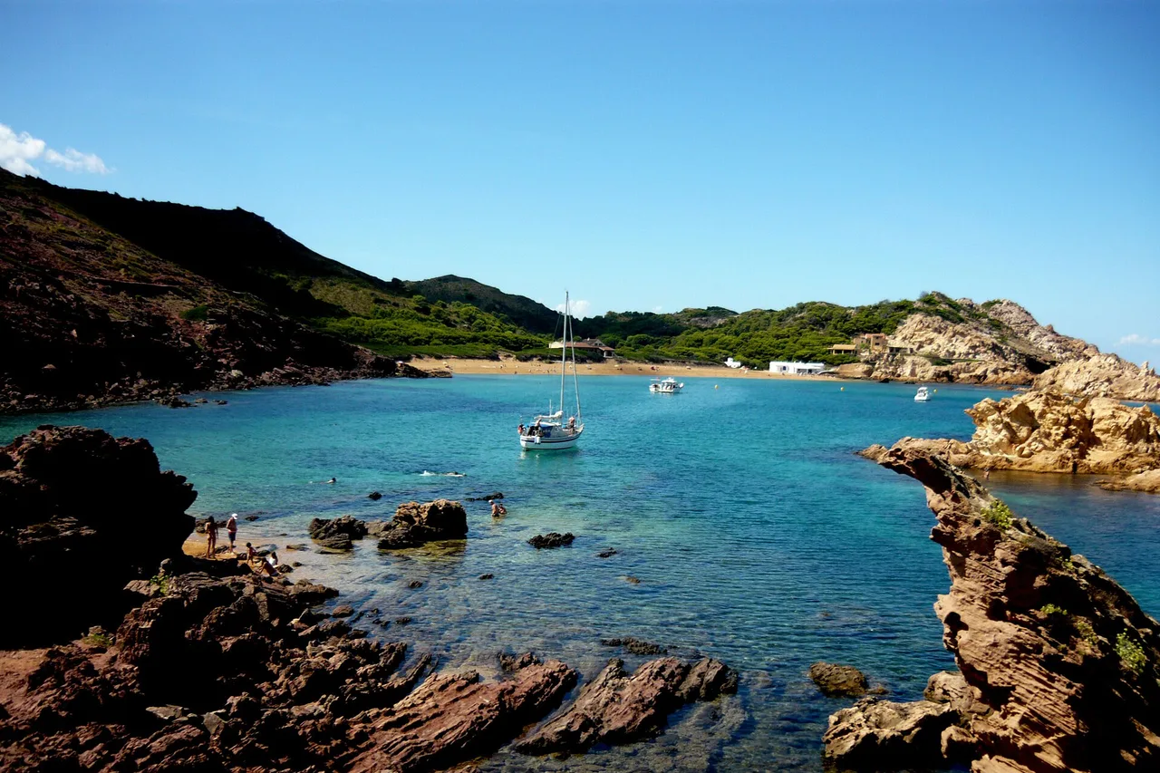 Cala Pregonda vue depuis les rochers : au premier plan des dalles de roche rousse presque rouge, l’eau bleue claire avec un voilier au mouillage et des nageurs, et au fond la plage de sable doré adossée à des collines basses.
