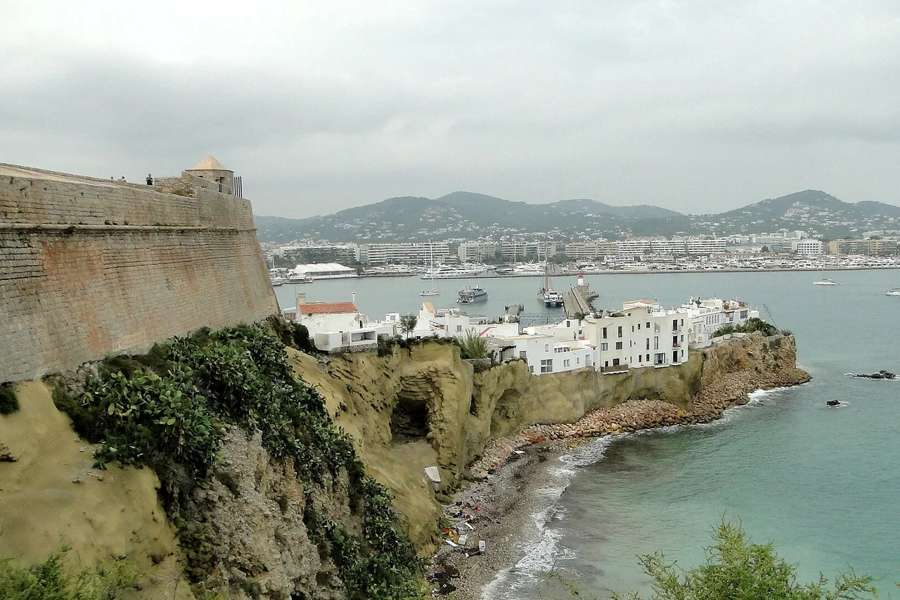 Die Mauern von Dalt Vila von einer Bastion aus: goldfarbenes Mauerwerk im Vordergrund, darunter die weißen Häuser des alten Hafens, türkisfarbenes Wasser und die Bucht von Eivissa mit ihren Hügeln im Hintergrund.