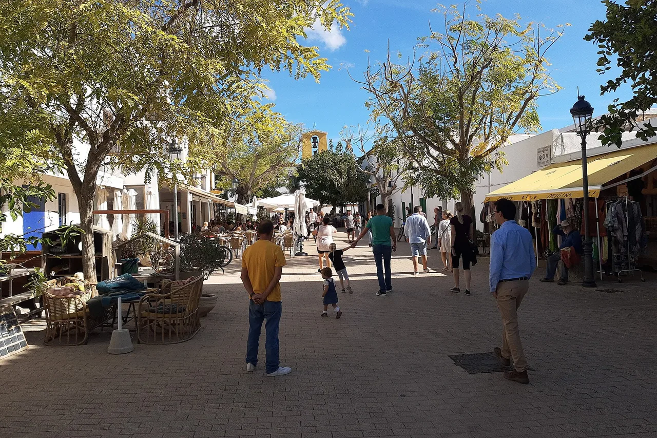 Der autofreie Dorfplatz von Santa Gertrudis de Fruitera auf Ibiza: niedrige weiß gekalkte Häuser, Café-Terrassen im Schatten der Bäume, Spaziergänger und Familien unter blauem Himmel.