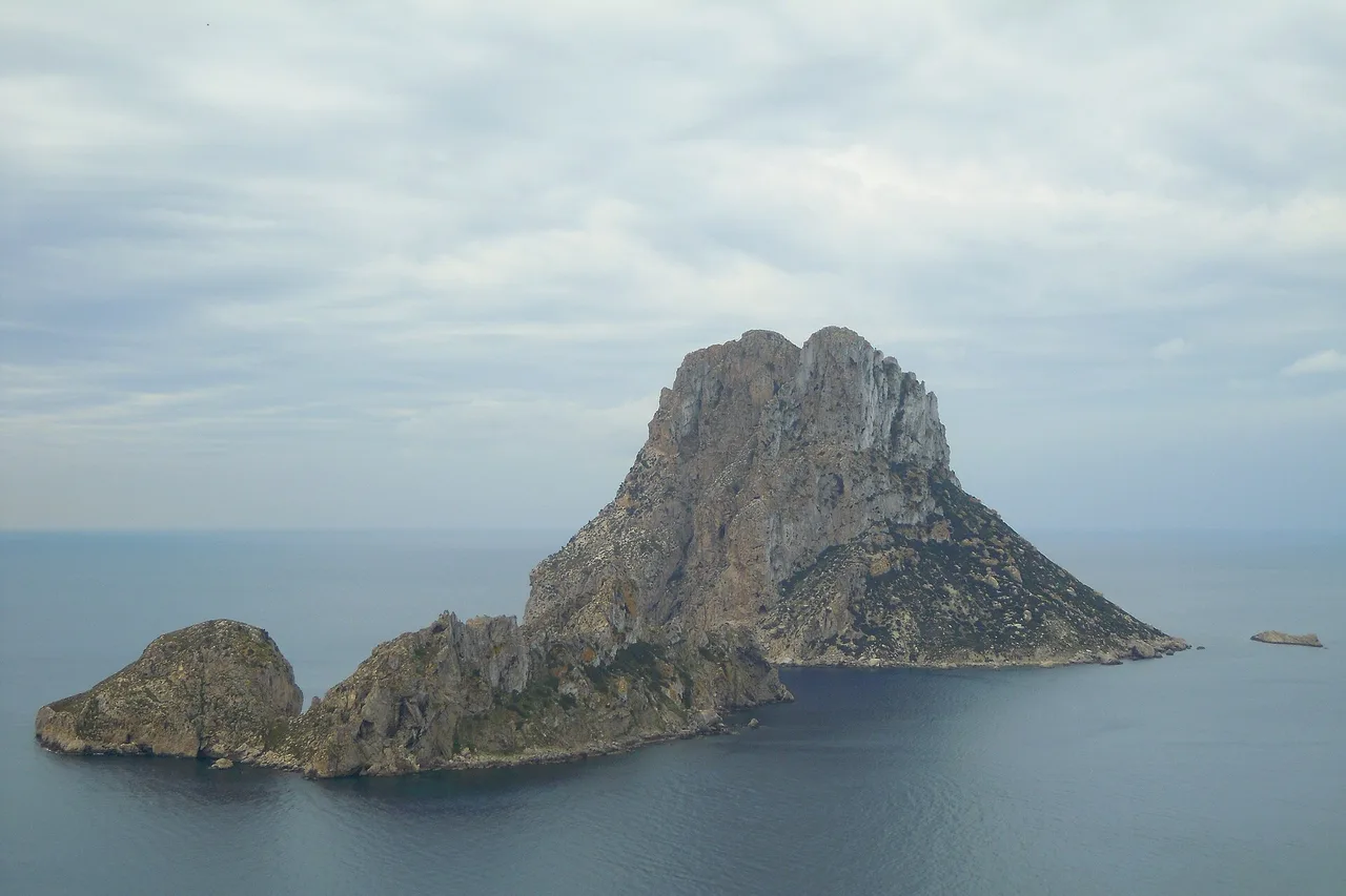 Die Felseninsel Es Vedrà erhebt sich aus einem ruhigen, silbrigen Meer vor Ibiza, davor der kleinere Nachbarfelsen Es Vedranell, darüber ein bewölkter Himmel.