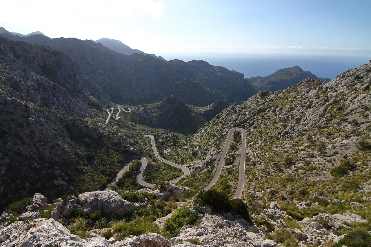 The Sa Calobra road seen from the Coll dels Reis: the ribbon of asphalt drops in tight hairpins into a limestone valley of the Serra de Tramuntana, two tiny cars on it, the sea on the horizon