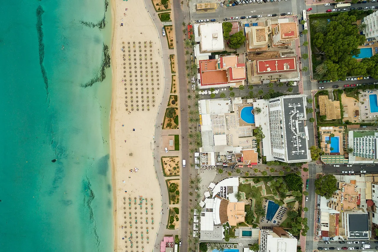 Vue aérienne verticale de la Platja de Palma : l’eau turquoise et le sable clair à gauche, les rangées de parasols, puis la première ligne d’hôtels avec leurs piscines bleues serrées les unes contre les autres.