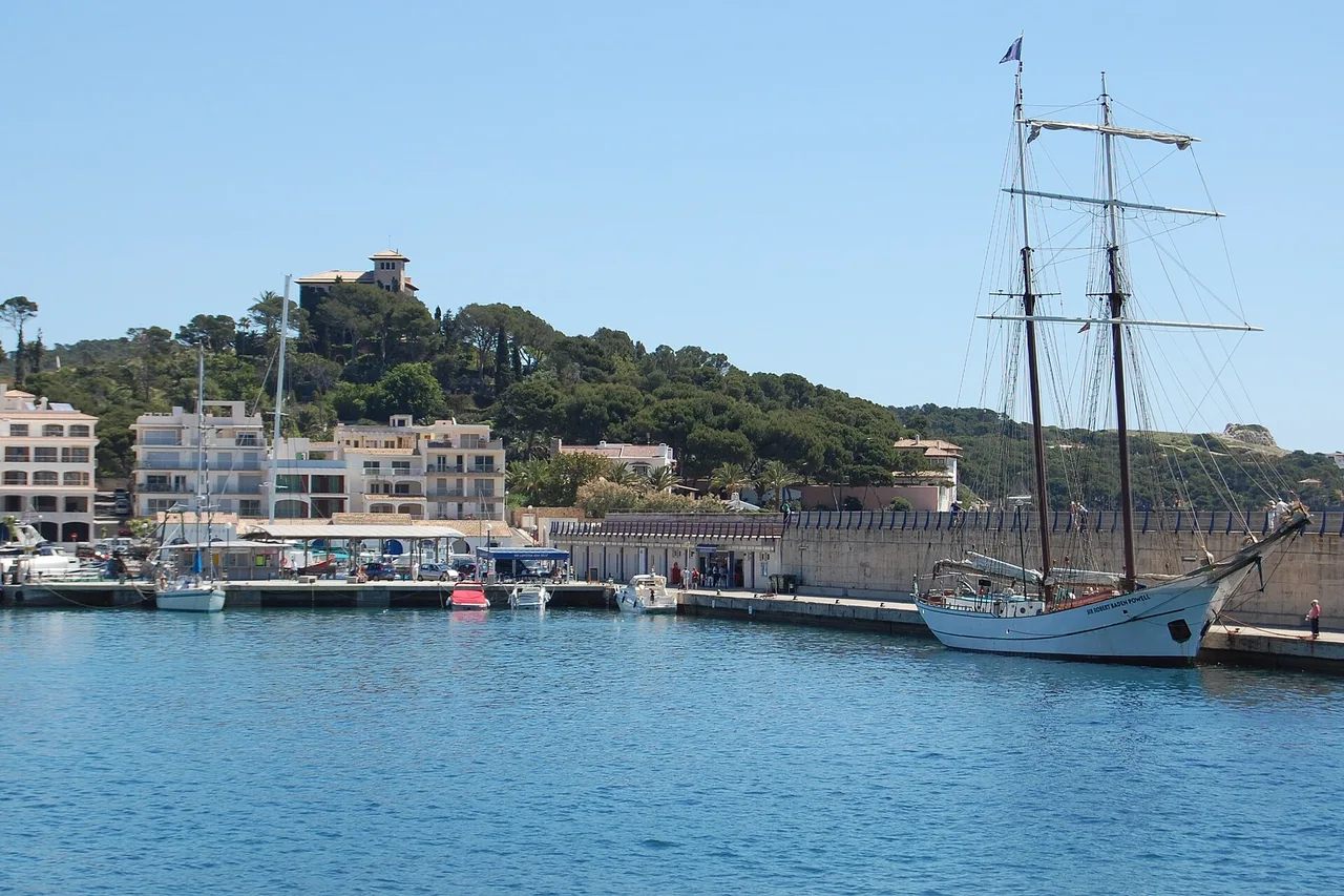 Le port de Cala Rajada : le quai, un grand voilier-école amarré, les immeubles blancs de la station derrière et la colline boisée coiffée d’une tour.