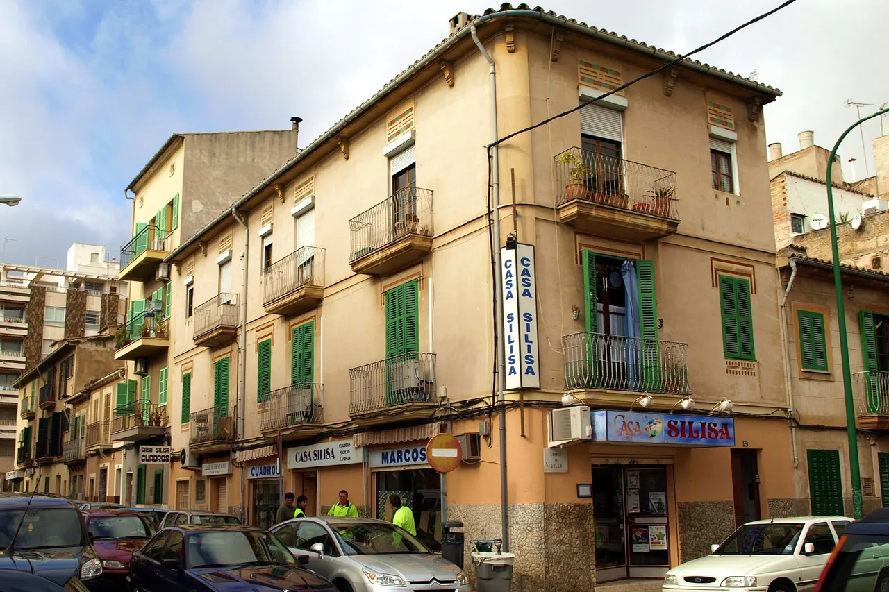 Un immeuble d’angle du quartier de Santa Catalina à Palma : pierre dorée, volets verts, balcons en fer, des commerces au rez-de-chaussée et des voitures garées le long du trottoir.