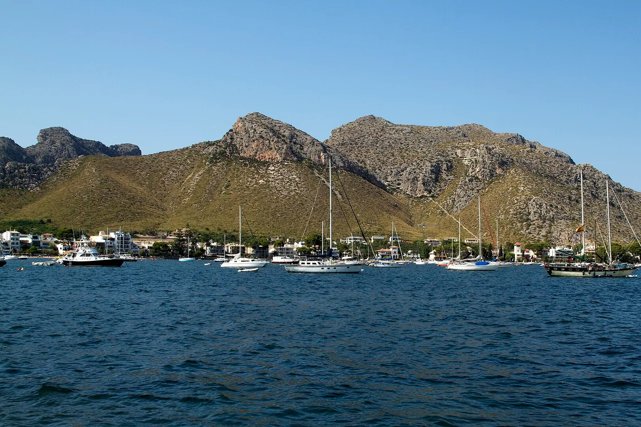 La baie de Port de Pollença vue de la mer : voiliers au mouillage, la ligne basse et blanche du village au fond, et derrière elle les reliefs secs de la péninsule.