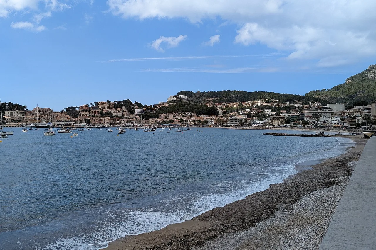 La baie du Port de Sóller vue depuis la plage de galets : l’anse presque fermée, les maisons basses du port alignées au pied de la montagne et les sommets de la Tramuntana derrière.