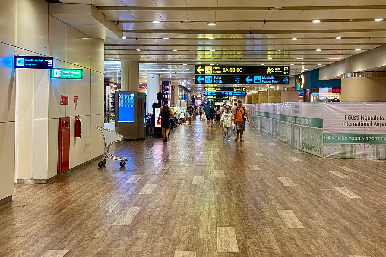 Ein Gang im internationalen Terminal des Flughafens Ngurah Rai mit Wegweisern zu den Gates, Toiletten und dem Gebetsraum, Reisende gehen mit Gepäckwagen
