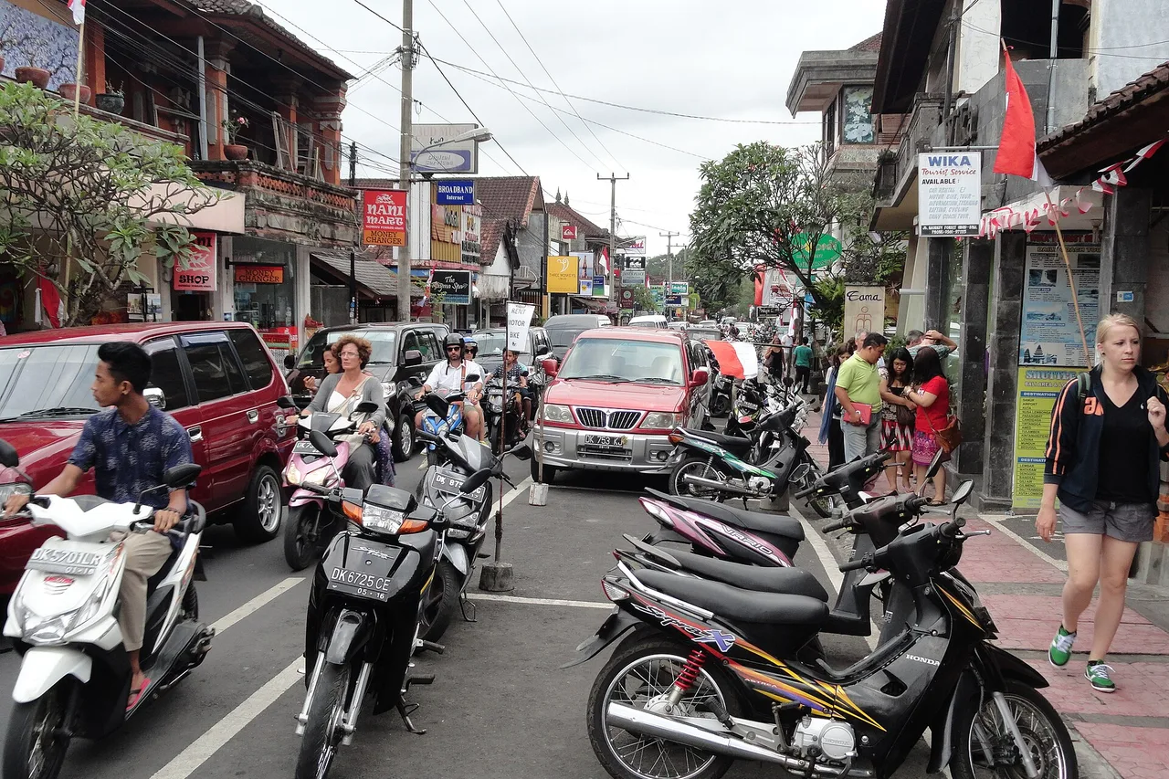 Eine enge Straße in Ubud mit Autos, in Doppelreihe geparkten Motorrollern am Rand und Fußgängern auf dem Gehweg