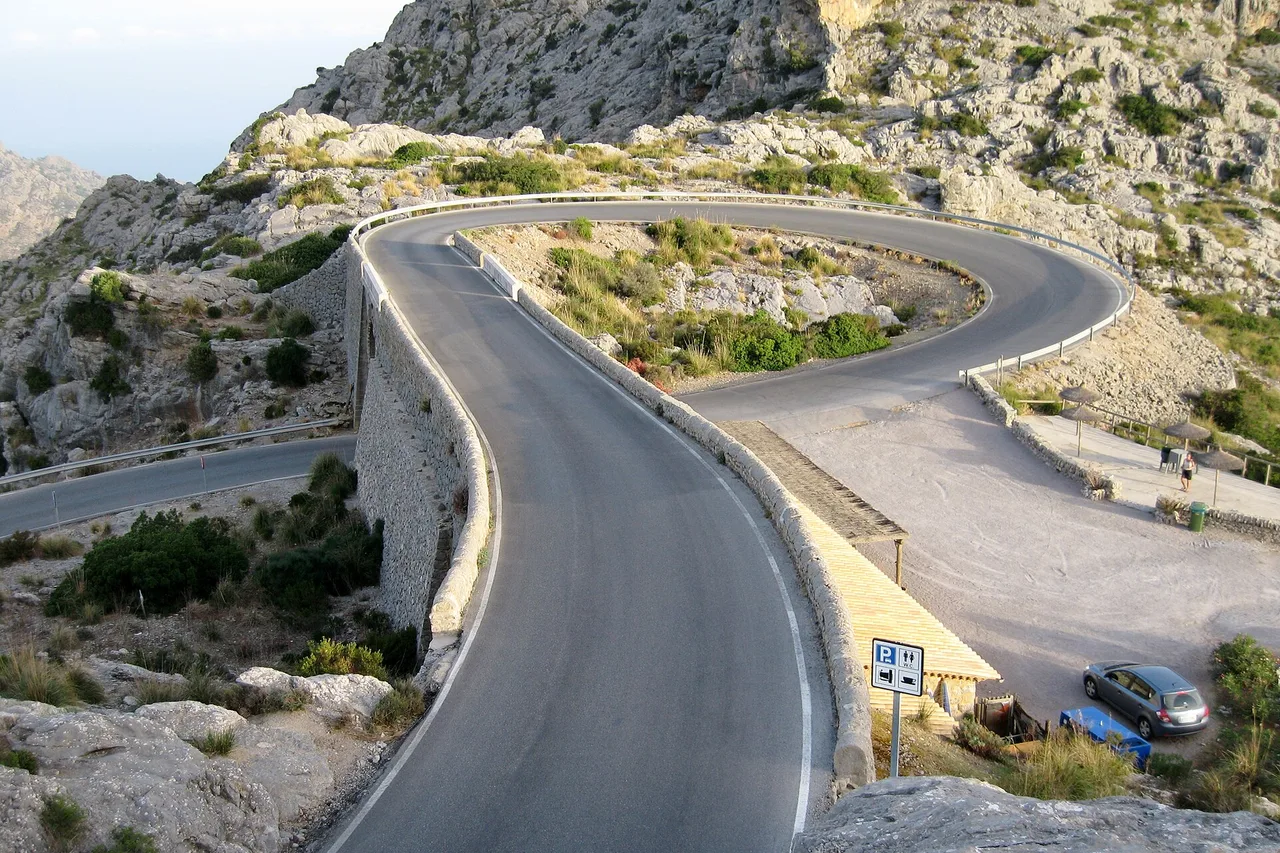 La curva Nus de sa Corbata sulla strada per Sa Calobra: una carreggiata con guard-rail gira attorno a un versante roccioso, con un piccolo parcheggio sulla destra