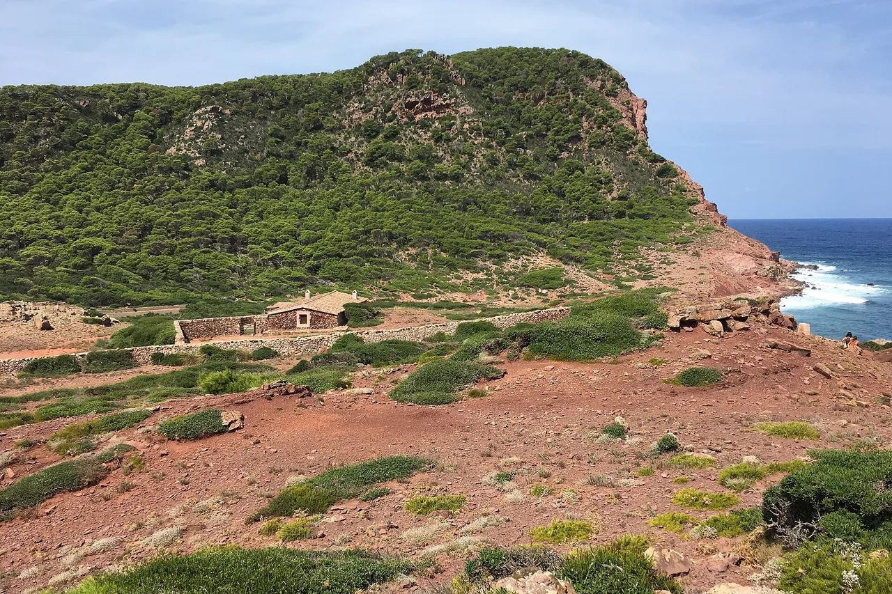 La côte nord de Minorque près de Cala Pilar : terre rouge, maquis ras et une maison de pierre isolée. Le Camí de Cavalls traverse ce paysage et fait le tour complet de l’île.