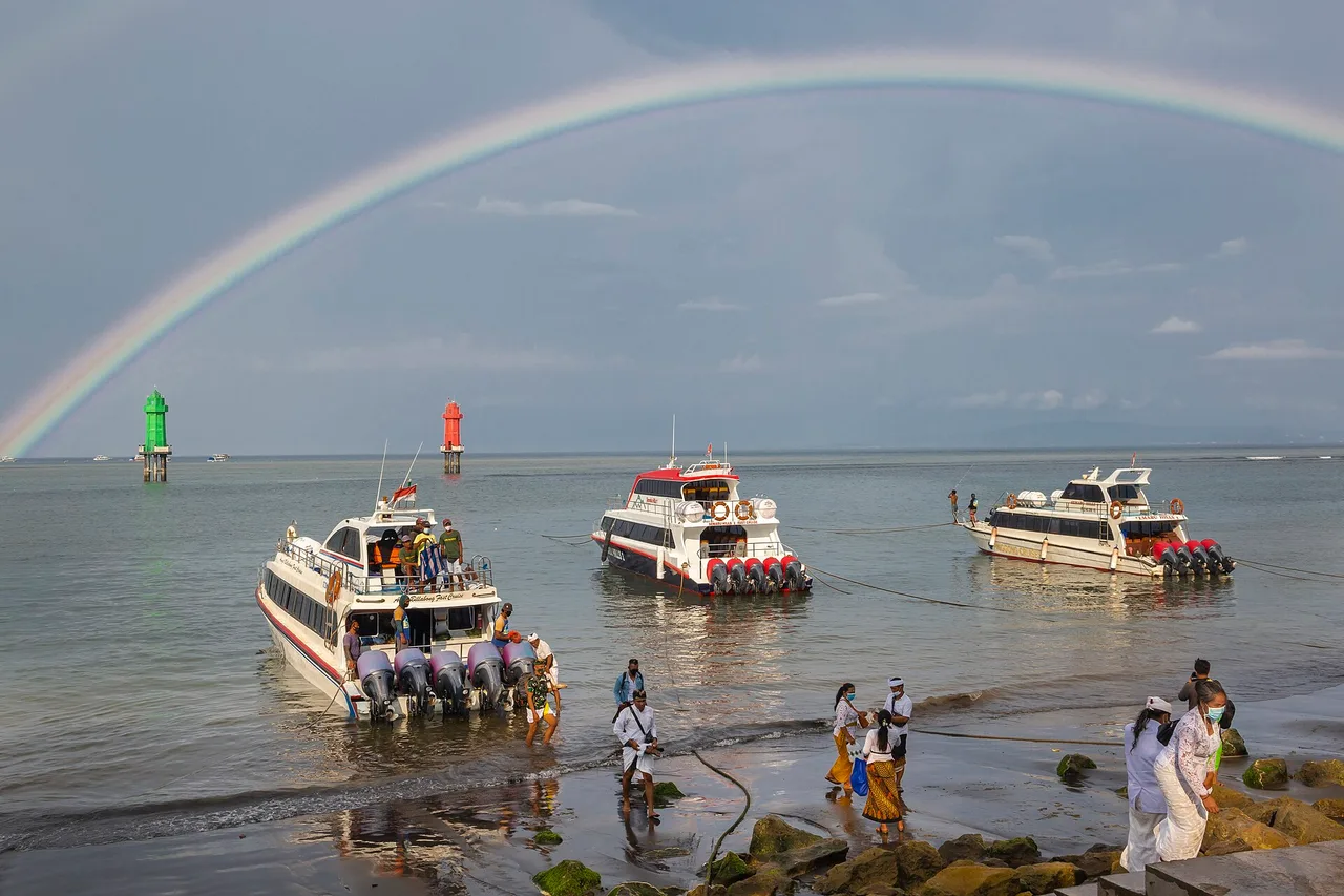 Several white fast boats pulled up in the shallow water of Sanur beach, people on the shore and a rainbow arching over the sea