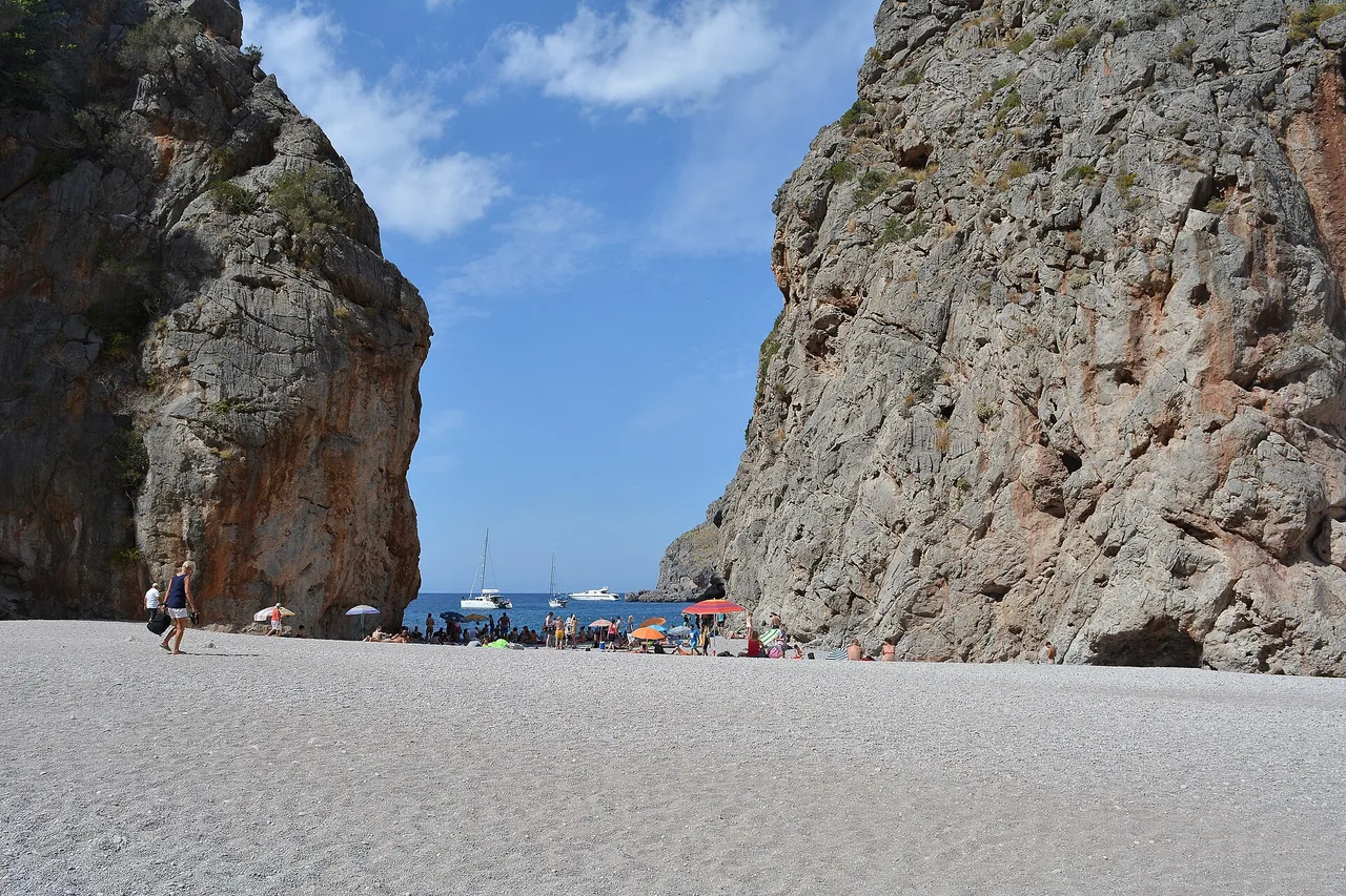Plage de galets de Sa Calobra entre deux hautes parois rocheuses, à l’embouchure du Torrent de Pareis, avec la mer et quelques parasols au fond