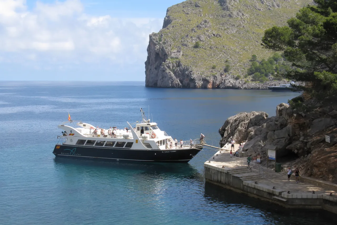 Un bateau d’excursion blanc chargé de passagers est amarré à un ponton dans une petite crique rocheuse de Sa Calobra