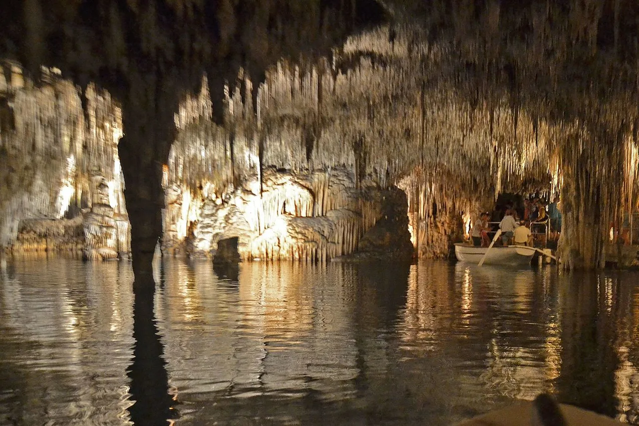 Salle éclairée des grottes du Drach avec stalactites, un lac aux reflets et une barque de visiteurs sur le bord