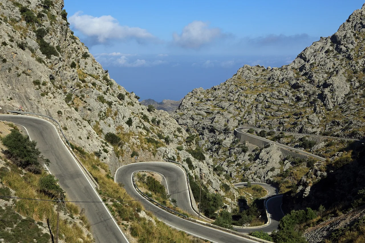 Une route en lacets aux virages en épingle descend dans une vallée rocheuse vers la côte, avec la mer et des nuages au fond