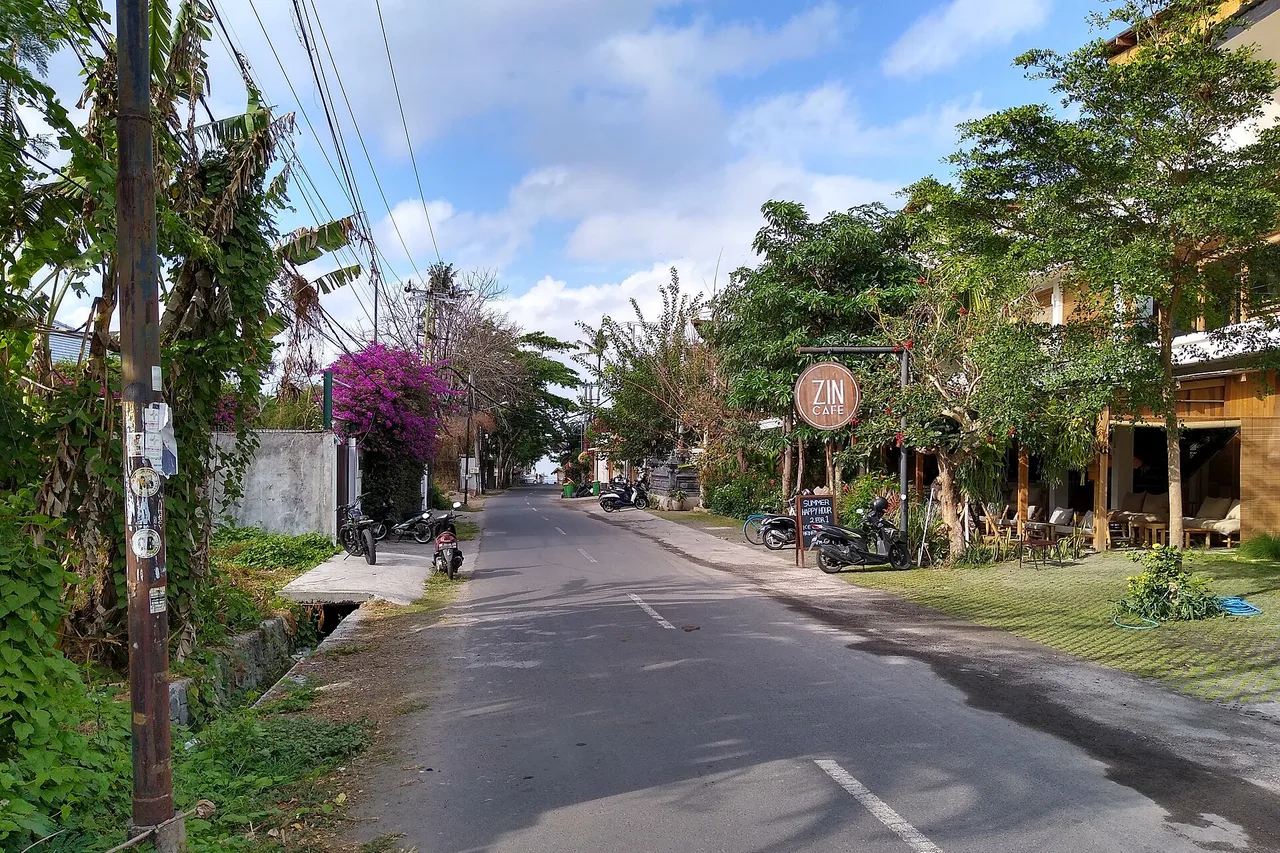 Rue étroite de Canggu avec un scooter, une enseigne de café et des bougainvillées sous un ciel bleu