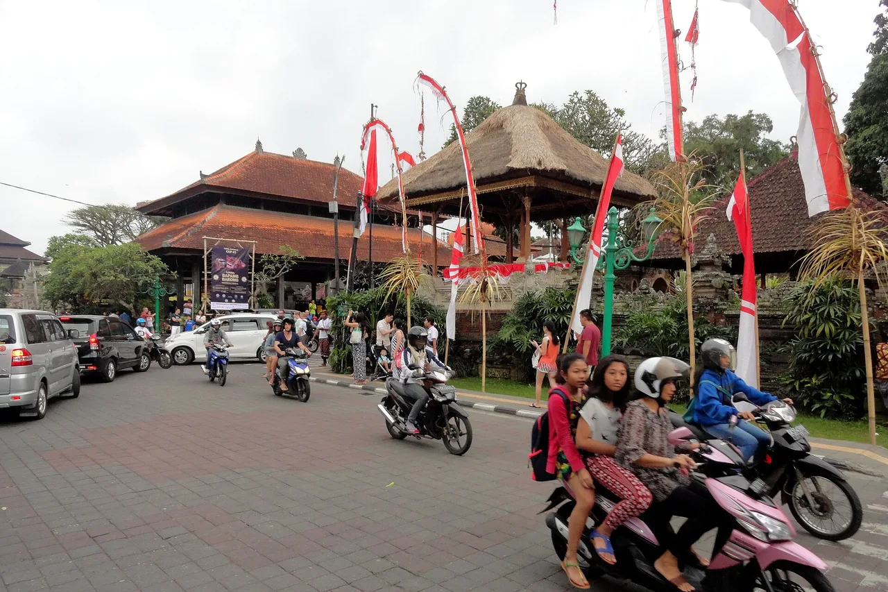 Scooters, voitures et piétons sur la Jalan Raya Ubud devant le palais d’Ubud, avec des penjor le long de la chaussée