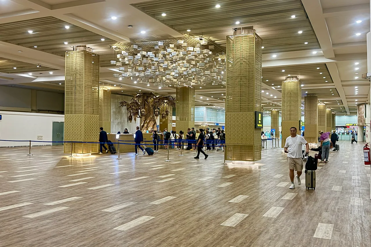 Ankunftshalle des internationalen Terminals von Ngurah Rai mit geflochtenen Säulen, großem Kronleuchter und Reisenden in der Schlange hinter blauen Absperrungen