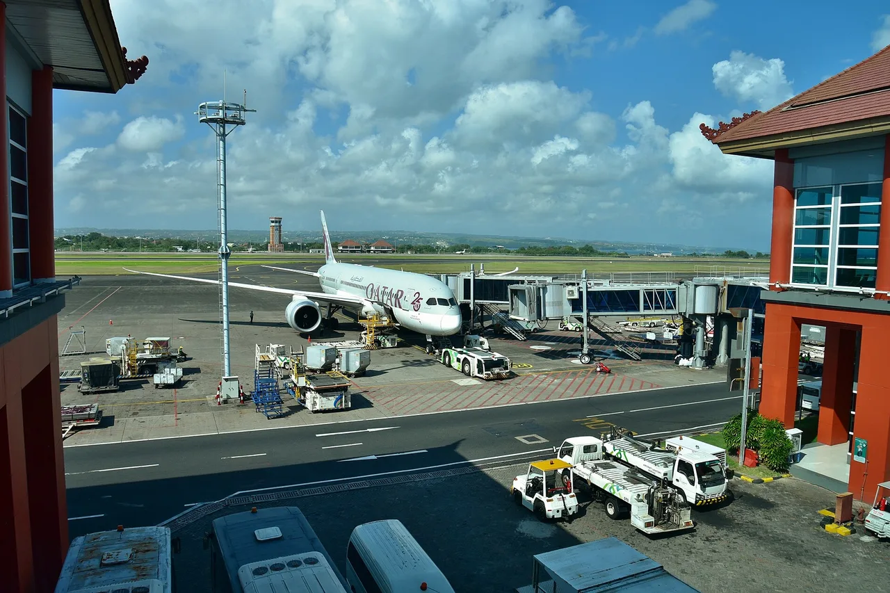 Eine Boeing 787 von Qatar Airways an der Fluggastbrücke des Flughafens Denpasar, davor Fahrzeuge auf dem Vorfeld und das Terminal mit rotem Dach