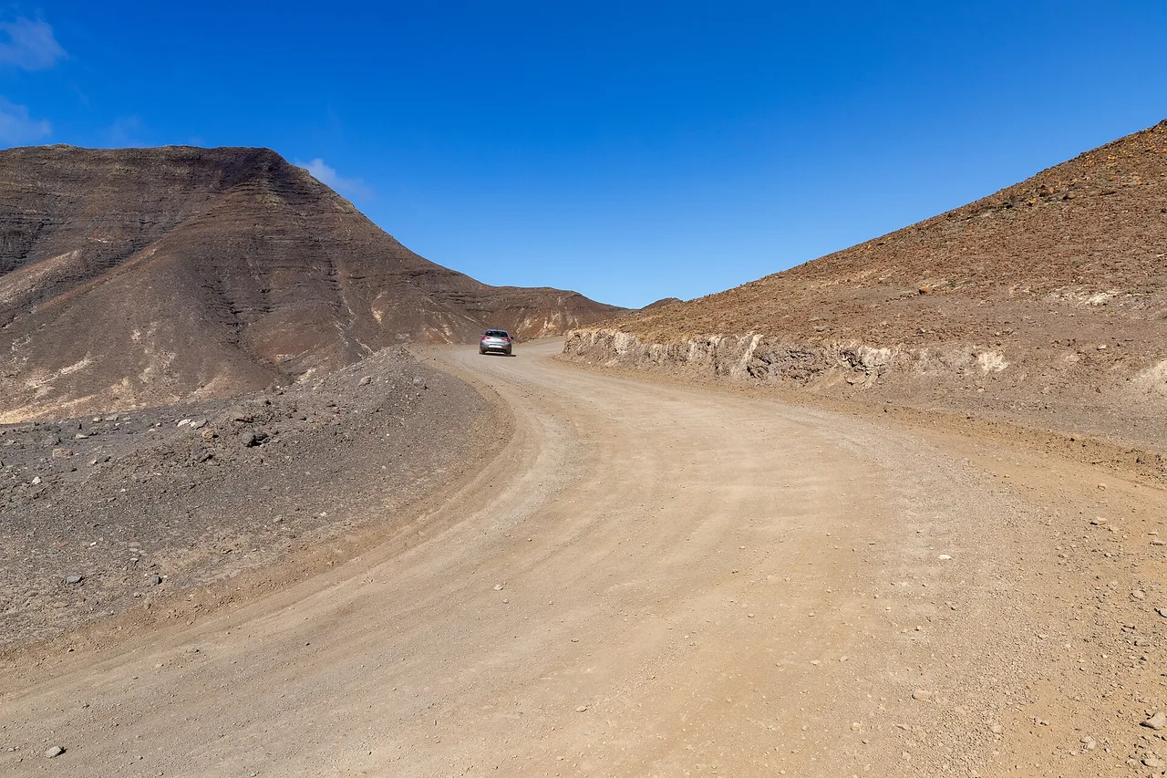 Una pista sterrata curva tra montagne brune a Fuerteventura, un’auto grigia procede in lontananza
