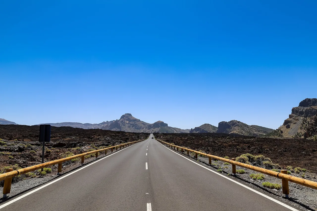 Una strada dritta con guardrail di legno attraversa un campo di lava scura nel Parco Nazionale del Teide, montagne all’orizzonte