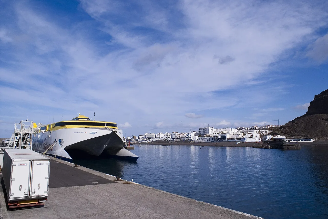 Il porto di Puerto de las Nieves ad Agaete: un catamarano di linea in banchina, dietro le case bianche del paese e una falesia scura.