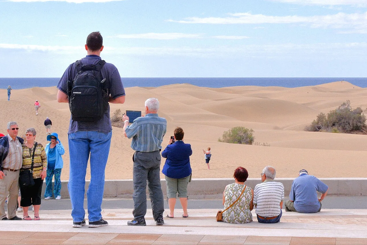 Visitatori in piedi o seduti sul bordo di una passeggiata guardano le dune di Maspalomas, con l'oceano all'orizzonte.