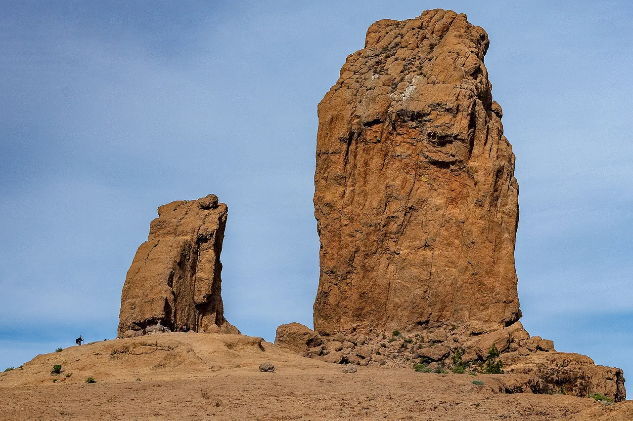 Il Roque Nublo, alto monolite di basalto bruno-rossastro, con una roccia più bassa alla sua sinistra; un escursionista minuscolo a sinistra dà la scala.