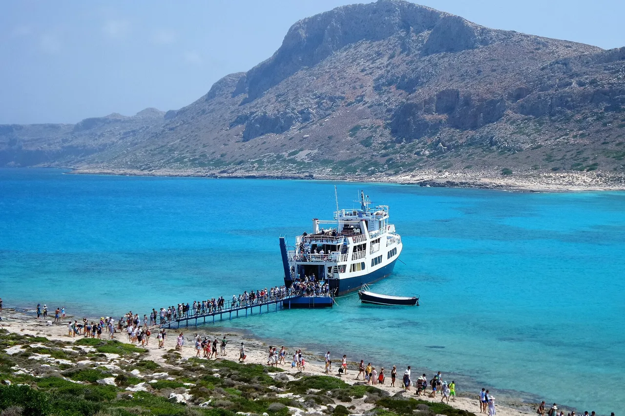 Una barca da escursione ormeggiata a un pontile sulla spiaggia di Balos, i visitatori sbarcano, sullo sfondo una montagna spoglia e acqua turchese