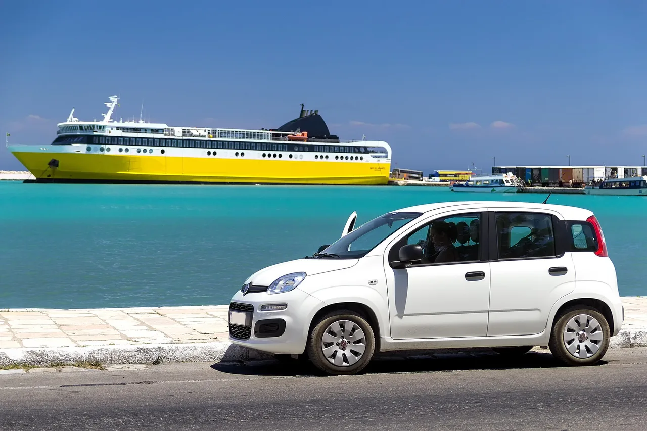 Una Fiat Panda bianca parcheggiata su una banchina del porto, con un traghetto giallo su acqua turchese alle spalle