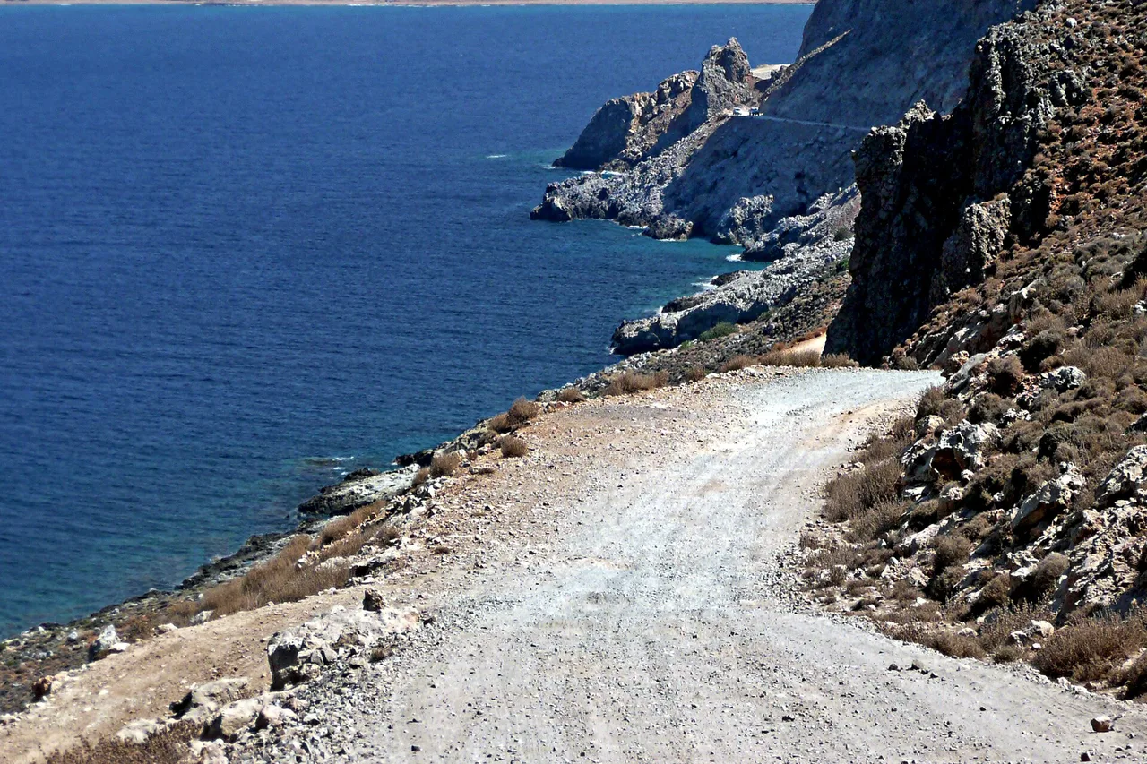Una pista di ghiaia chiara costeggia una scogliera rocciosa, con il mare blu intenso a sinistra