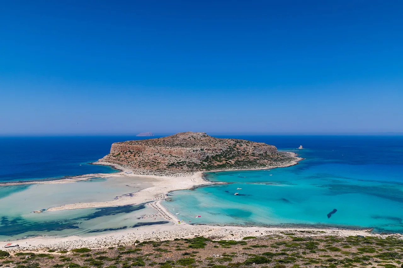 Vista aérea de la laguna de Balos: agua turquesa poco profunda y bancos de arena clara, un promontorio rocoso en el centro y detrás el mar azul intenso