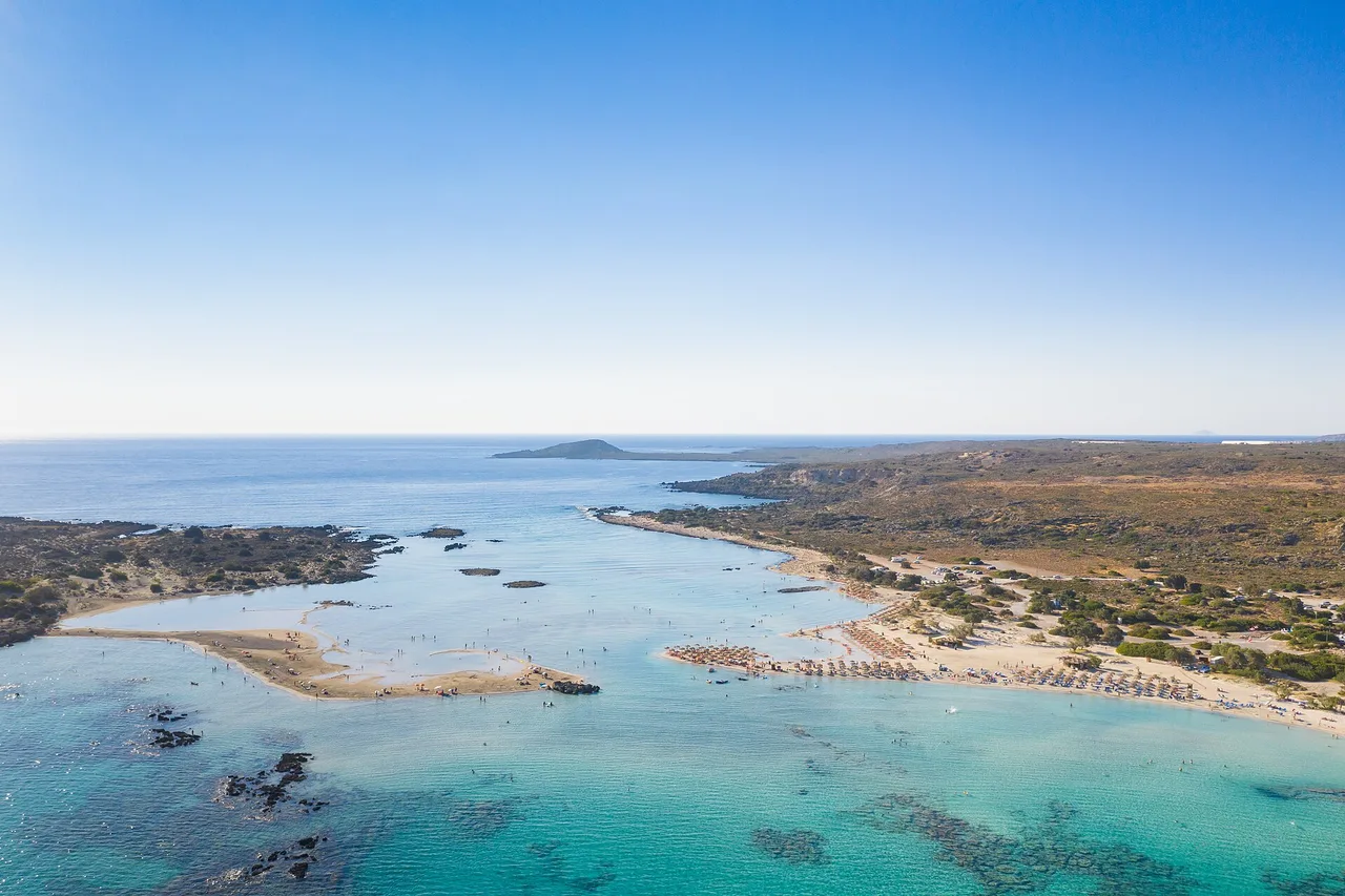 Vista aérea de una laguna turquesa poco profunda con un banco de arena, a la derecha una playa con sombrillas y en el horizonte una pequeña isla