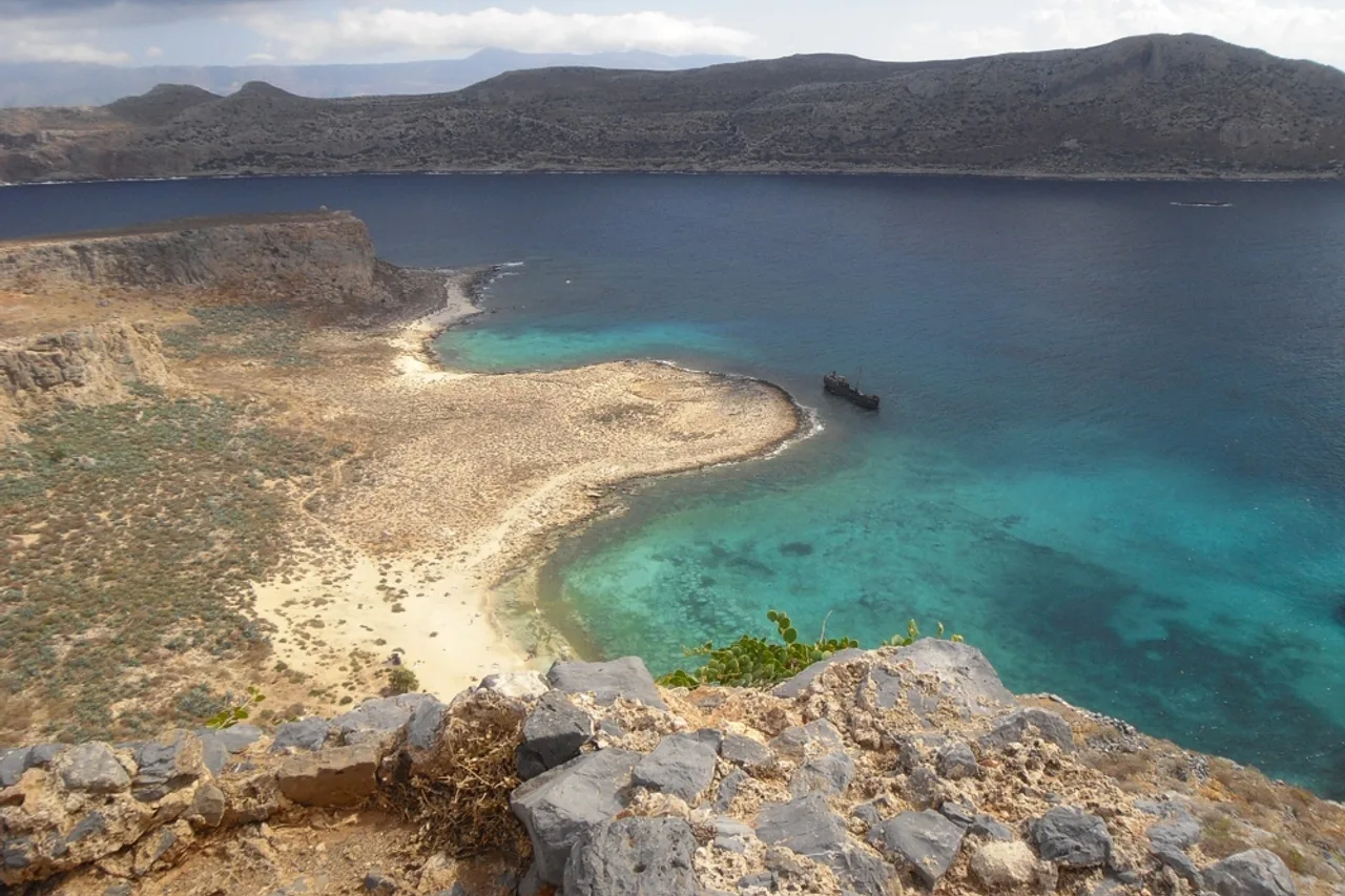 Vista desde un muro de piedra sobre una lengua de arena y una bahía turquesa en Gramvousa, con un naufragio oscuro varado en la orilla