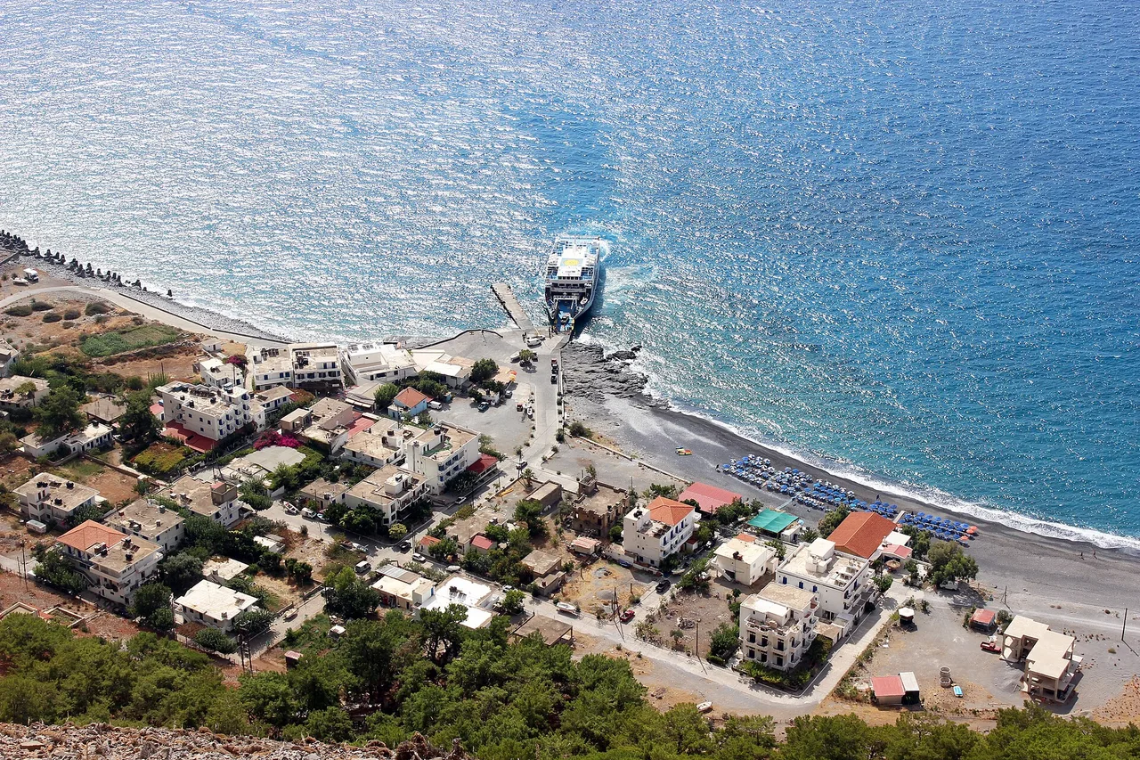 Vista desde arriba del pueblo costero de Agia Roumeli: casas blancas, un ferri en el muelle y una playa de guijarros con tumbonas azules