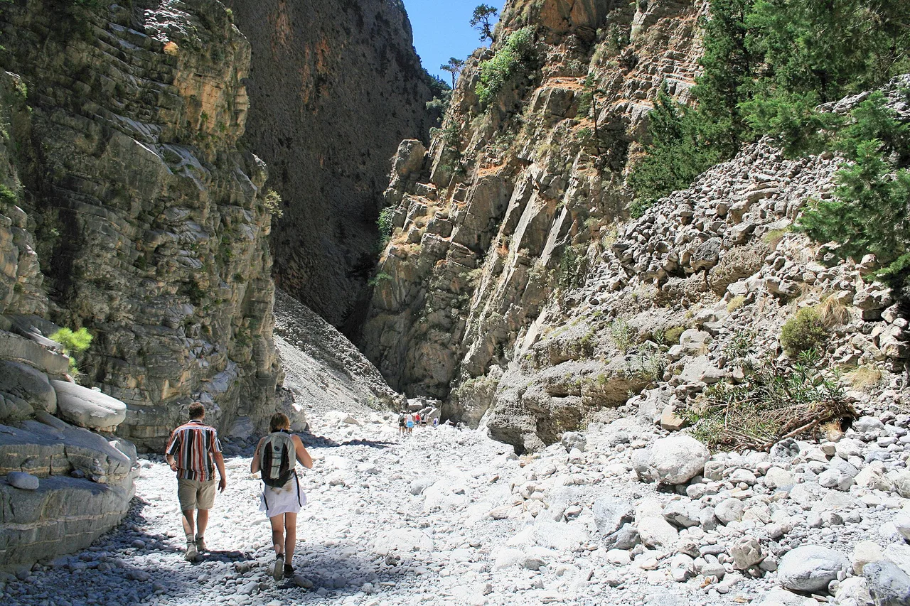 Dos excursionistas caminan por un sendero de piedras claras entre las paredes abruptas de una garganta