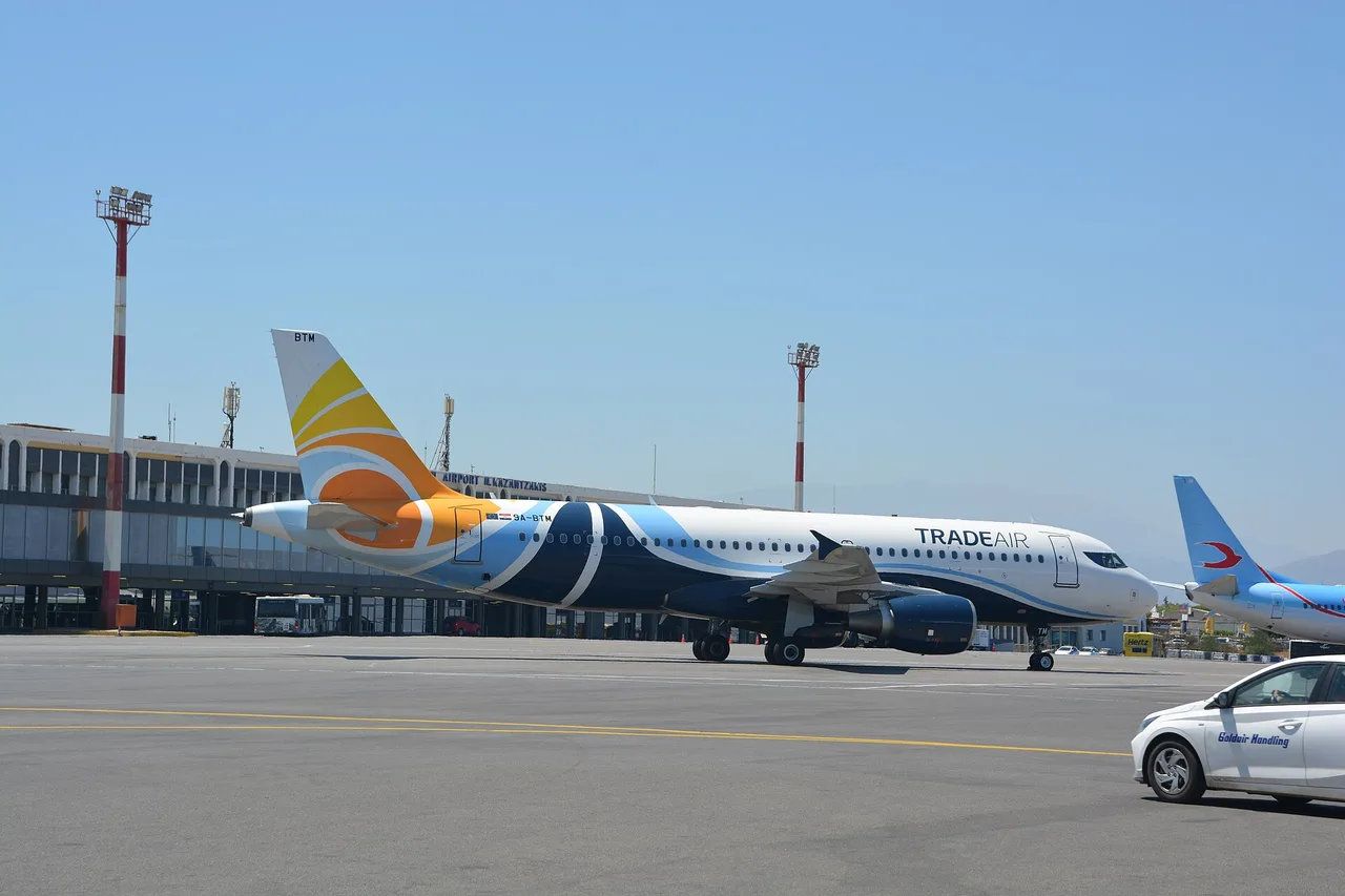 Das Terminal des Flughafens Heraklion mit der Aufschrift Airport N. Kazantzakis, davor ein Airbus von Trade Air auf dem Vorfeld und rot-weiße Lichtmasten