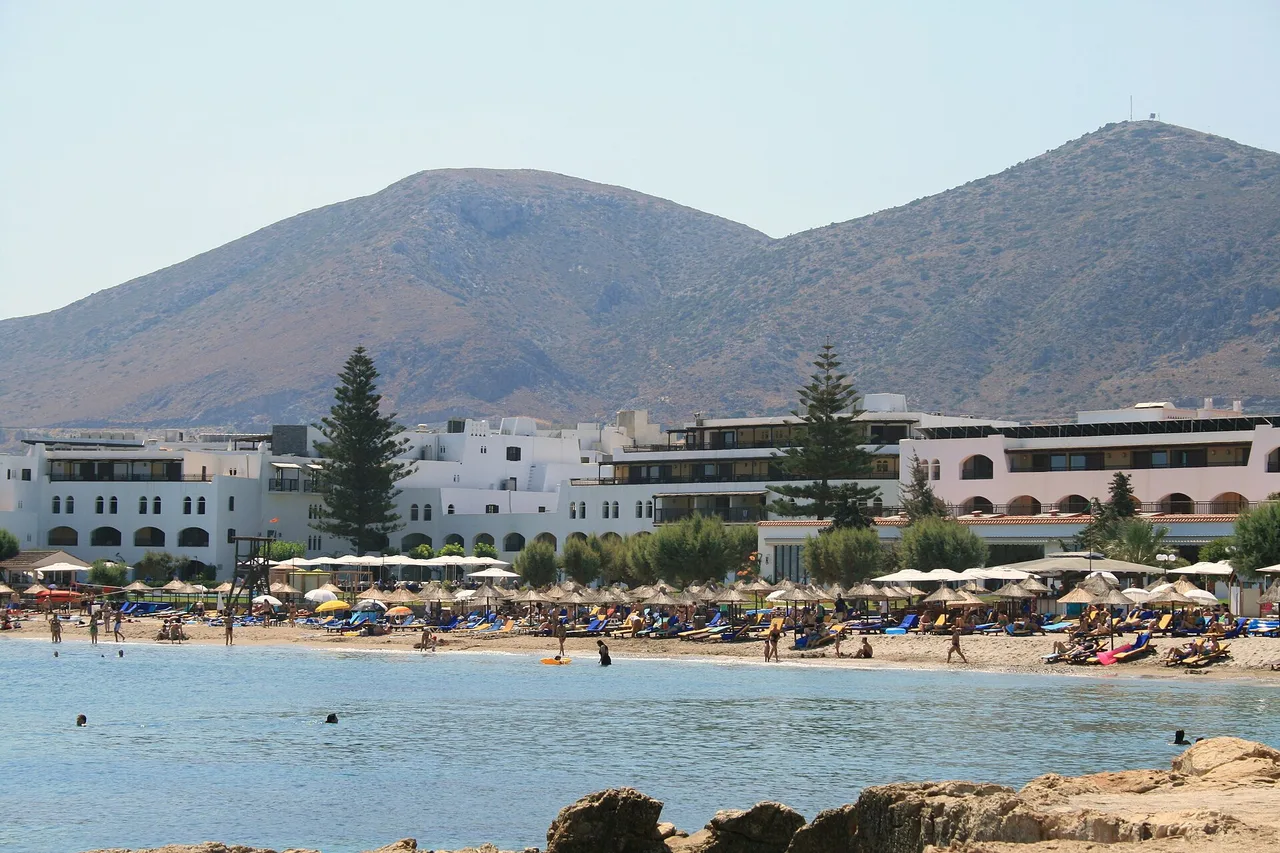 Sandstrand mit Sonnenschirmen vor weißen Hotelbauten am Fuß kahler Hügel in Hersonissos auf Kreta