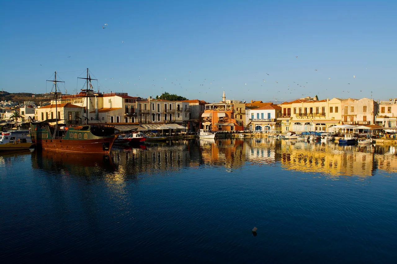 Der venezianische Hafen von Rethymno im Morgenlicht, bunte Fassaden spiegeln sich im Wasser, ein zweimastiges Holzschiff liegt am Kai