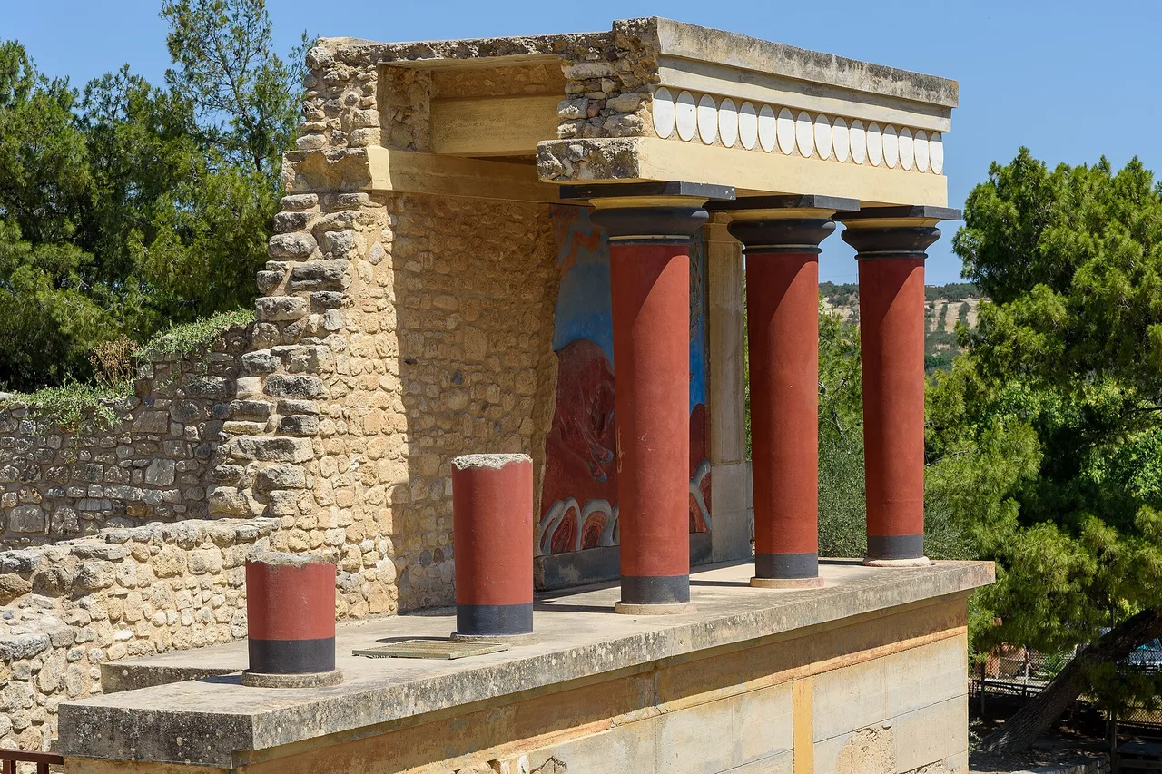 L’ingresso nord del palazzo di Cnosso, a Creta: colonne rosse ricostruite sotto un tetto di pietra, con pini e cielo azzurro dietro.