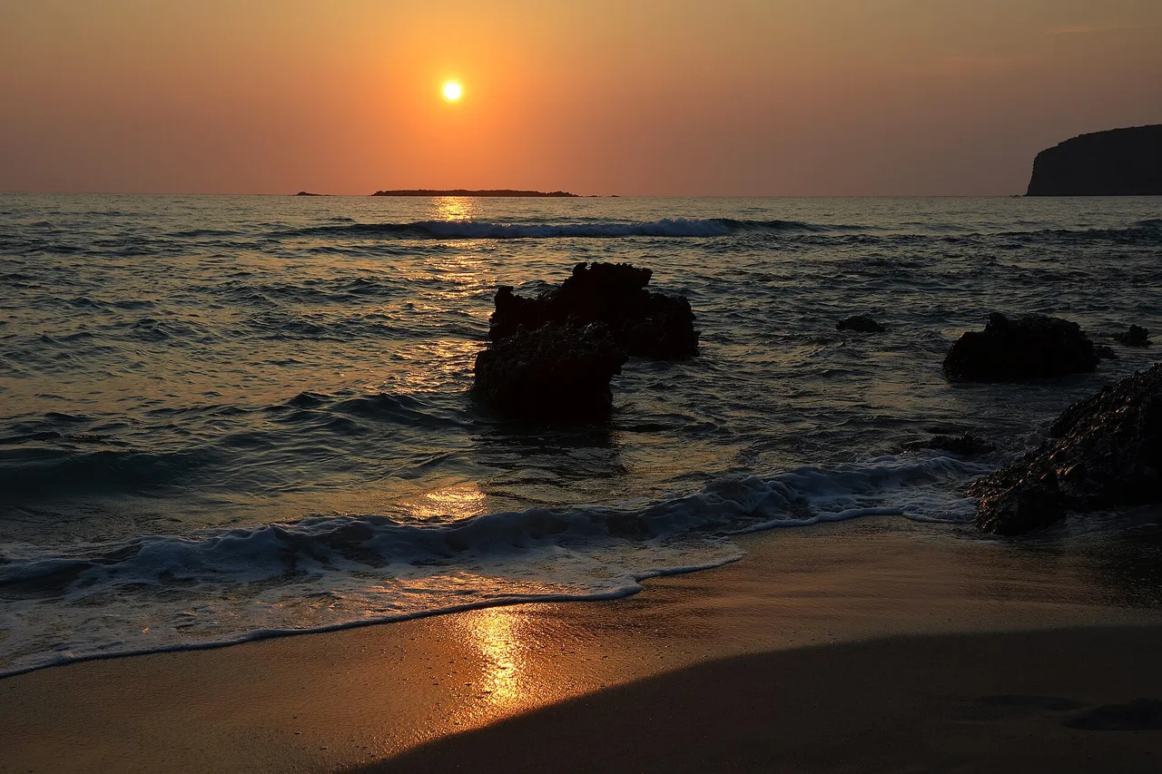 Coucher de soleil sur la mer à Falassarna, à la pointe ouest de la Crète : le disque orange touche l’horizon et sa lumière court sur le sable mouillé, entre des rochers noirs pris dans le ressac.