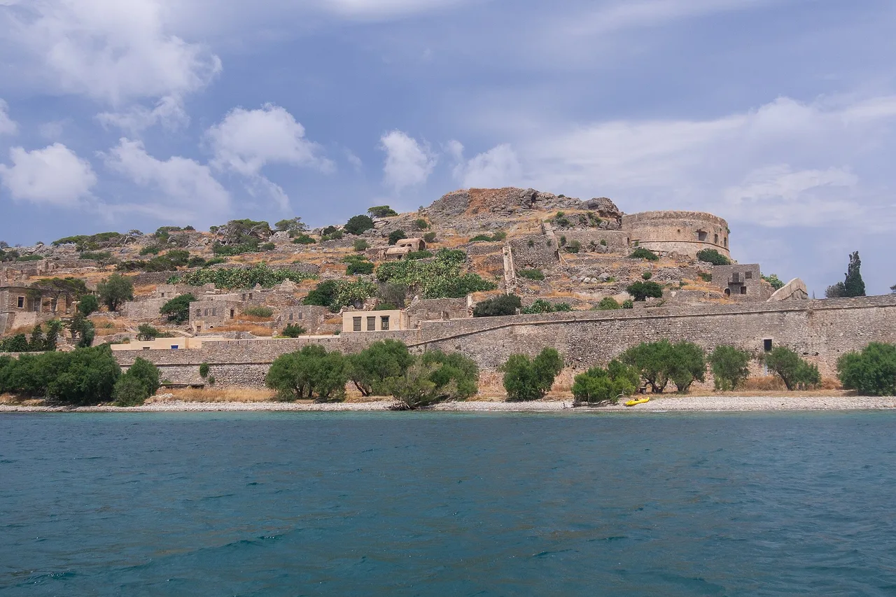L’îlot fortifié de Spinalonga vu depuis un bateau : remparts vénitiens, maisons en ruine étagées sur la pente pelée, mer turquoise au premier plan et ciel nuageux.