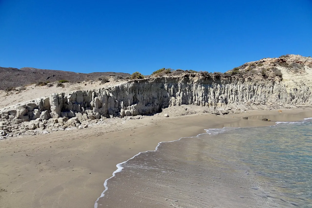 Une plage déserte du sud-est de la Crète : sable clair, vaguelettes, une barre de roche blanche stratifiée au bord de l’eau et des collines arides sans la moindre construction sous un ciel bleu vif.