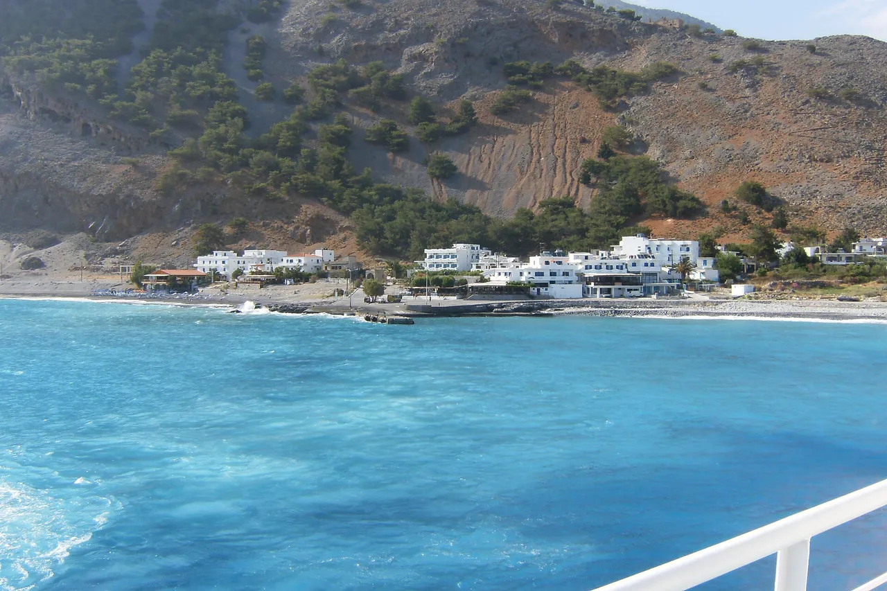 El pueblo de Agia Ruméli visto desde la cubierta de un barco, barandilla blanca en primer plano, casas blancas alineadas entre el mar turquesa y la montaña árida.